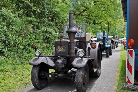 Ein alter Lanz Bulldog Traktor fährt auf einem schmalen Weg durch eine grüne Landschaft. Im Hintergrund sind weitere Traktoren zu sehen.