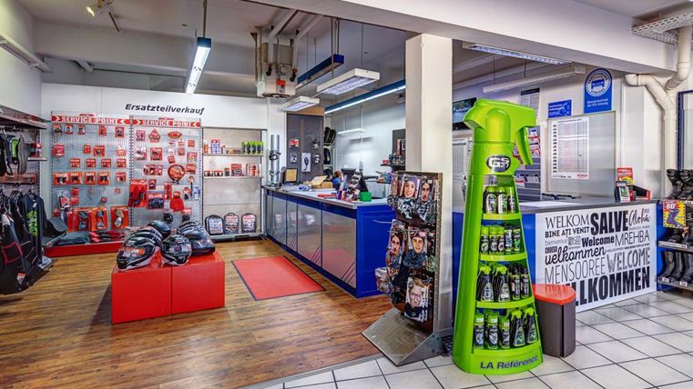 Ein Verkaufsraum mit einem Empfangstresen und verschiedenen Produkten an den Wänden. Es gibt Regale mit Zubehör, und der Boden ist mit Fliesen und Holz ausgestattet.