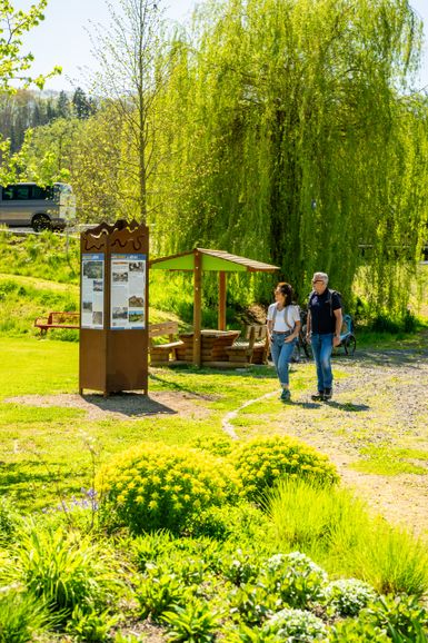 In einer sonnigen, grünen Naturlandschaft steht links eine Informationstafel aus Holz. Daneben verläuft ein heller Weg über eine kleine Holzbrücke. Zwei Personen befinden sich auf dem Weg in der Nähe der Tafel. Umgeben ist die Szene von Wiesen, Sträuchern und Bäumen in kräftigen Grüntönen. Die Aufnahme zeigt einen ruhig gelegenen Rast- und Informationspunkt entlang eines Wanderwegs