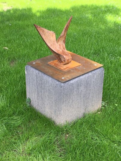 Une sculpture moderne d'un oiseau en métal se dresse sur un socle gris. Elle se trouve sur une prairie verte.