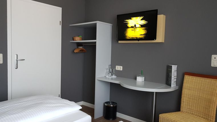 A modern hotel room with two beds, a television on the wall, and a small desk. The walls are gray and there is a rattan chair.