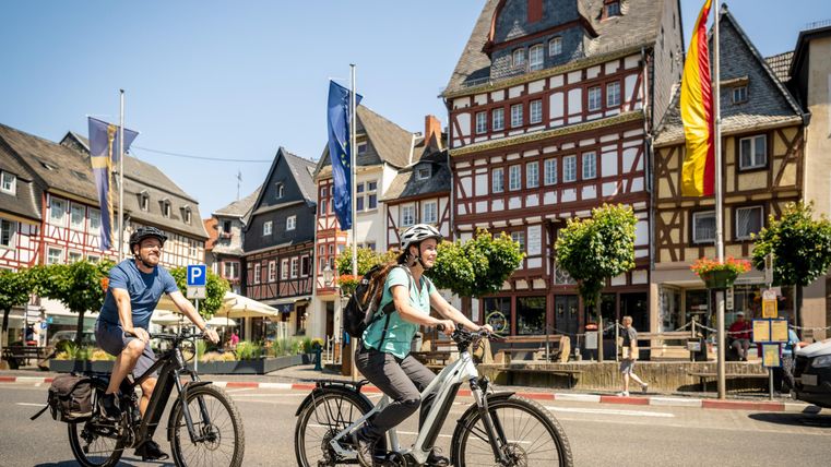 Zwei Radfahrer fahren auf einer Straße in einer malerischen Stadt. Im Hintergrund sind typisch deutsche Fachwerkhäuser und Bäume zu sehen.