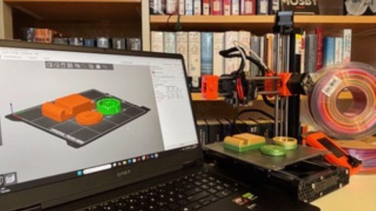 Ein Laptop zeigt eine 3D-Design-Software, während ein 3D-Drucker auf einem Tisch arbeitet. Im Hintergrund sind Bücherregale mit verschiedenen Büchern zu sehen.