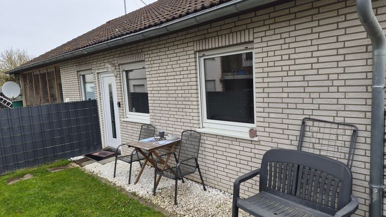 A simple house with a paved terrace and garden furniture. In the background, a gray wall is visible.