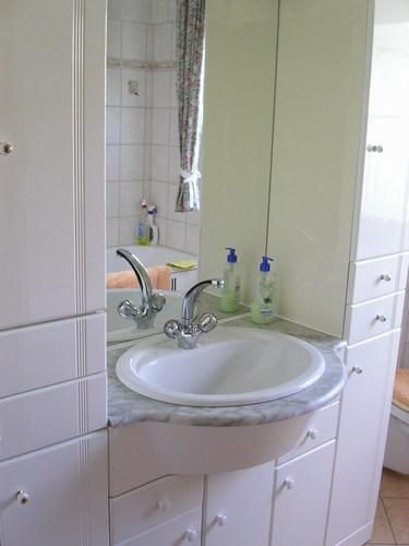 Een lichte badkamer met een ovaal fonteintje en een grote spiegel. De muren zijn betegeld en er staan enkele verzorgingsproducten op de plank.