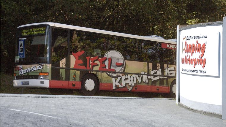 A bus with colorful graffiti is standing by a path near a campsite. Trees can be seen in the background.