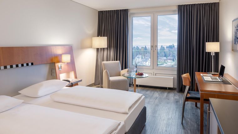 Een modern hotelkamer met twee bedden en een gezellige werkplek. Grote ramen bieden uitzicht op de stad en zorgen voor veel natuurlijk licht.