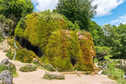 Mos bedekte waterval bij Nohn, omringd door weelderig groen. Een klein pad loopt langs het water. De lucht is blauw met enkele wolken.