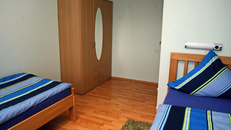 Een eenvoudige kamer met twee bedden en een kast. De vloer is van hout en er ligt een klein tapijt.