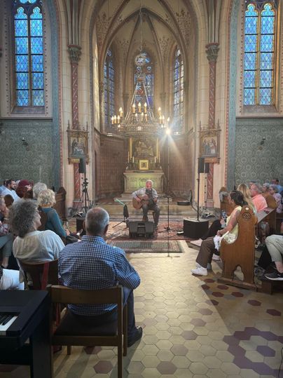 A musician plays in a church for an audience. The atmosphere is warm and inviting with beautiful windows and historic architecture.