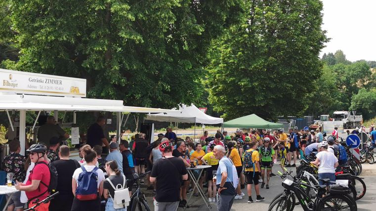 Eine Gruppe von Radfahrern steht an einem Verkaufsstand und wartet auf Essen und Getränke. Bäume und Zelte im Hintergrund schaffen eine entspannte Atmosphäre.