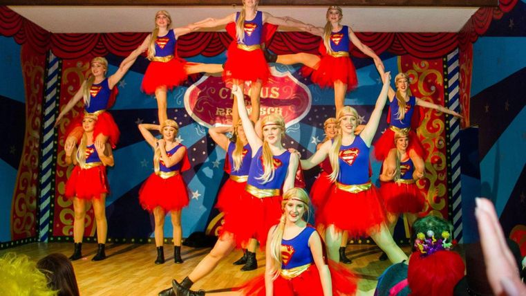 Een groep dansers in rode tutu's en blauwe tops voert een voorstelling op een podium uit. Ze tonen indrukwekkende acrobatische figuren in een feestelijke omgeving.