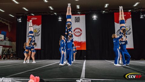 Eine Cheerleading-Gruppe führt beeindruckende Stunts in blauen Uniformen auf. Die Athleten zeigen ihr Können auf einer Wettkampfbühne mit unterstützenden Bannern im Hintergrund.