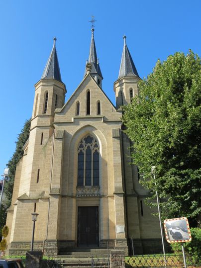 An impressive church with three towers and a high facade. The clear blue sky frames the building and the surrounding trees.