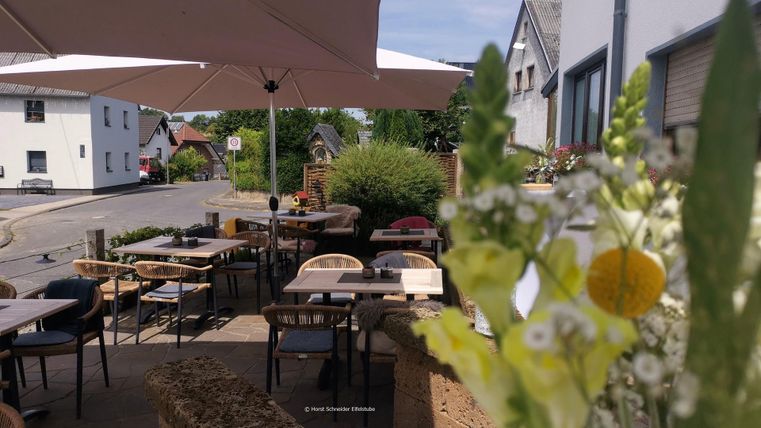 Ein gemütlicher Außenbereich eines Cafés mit Tischen und Stühlen. Sonnenschirme bieten Schatten und Blumen schmücken den Tisch.
