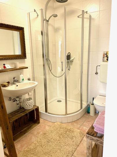 A modern bathroom with a shower and a sink. The walls are light-colored tiles and there is a large mirror above the sink.