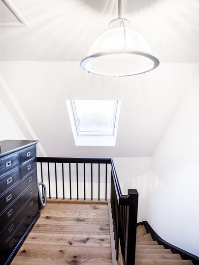 Ein heller Treppenaufgang mit Holzfußboden und einem großen Dachfenster. Zu sehen sind ein moderner Lampenschirm und eine schwarze Kommode.