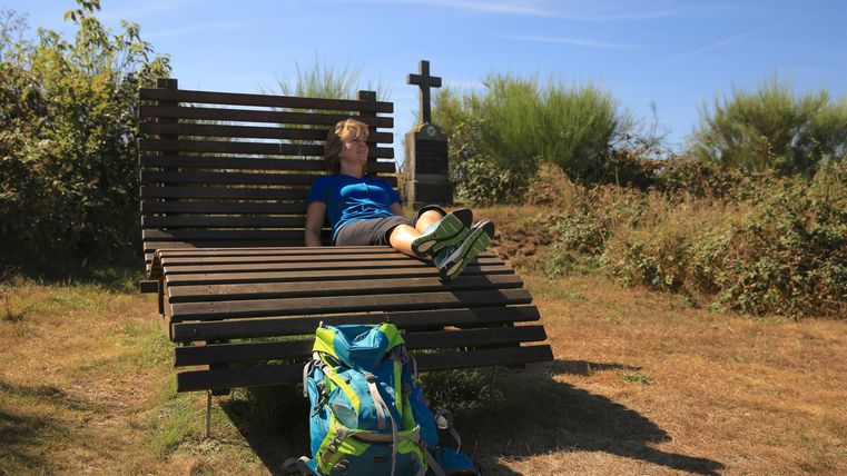 Eine Person entspannend auf einer Holzliege in der Natur. Im Hintergrund ist ein Kreuz und ein Rucksack zu sehen.