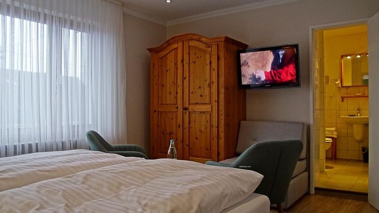 Une chambre confortable avec deux lits, une armoire en bois et une télévision. La fenêtre est décorée de rideaux blancs et laisse entrer la lumière naturelle.