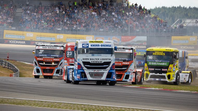 Renntrucks fahren auf einer Rennstrecke. Im Hintergrund sind Zuschauer zu sehen.