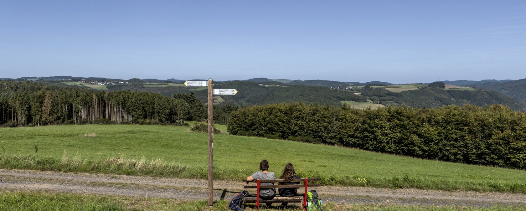 Zwei Personen sitzen auf einer Bank und blicken auf eine grüne Landschaft mit Hügeln und Wäldern. Ein Wegweiser steht daneben., © TI Hocheifel-Nürburgirng©BaumannFotografie