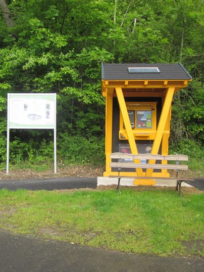 Eine gelbe Informationsstelle steht in einer grünen Landschaft. Daneben befindet sich ein Schild mit Informationen.