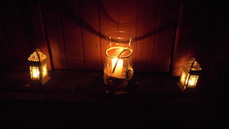 Eine ruhige und stimmungsvolle Szenerie mit einer brennenden Kerze in einem Glas und zwei leuchtenden Laternen. Das Licht schafft eine gemütliche Atmosphäre.