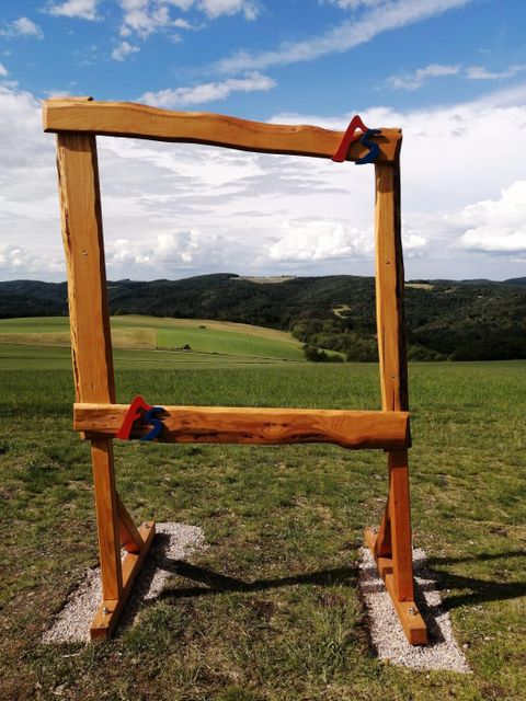Een houten frame staat op een weiland met uitzicht op zachte heuvels en een bewolkte lucht. Het landschap lijkt vredig en uitnodigend.