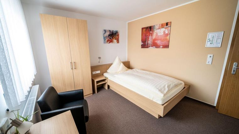 Een eenvoudige kamer met een eenpersoonsbed, een bureau en een stoel. De muren zijn in neutrale kleuren geschilderd, en er zijn een paar decoratieve foto's.