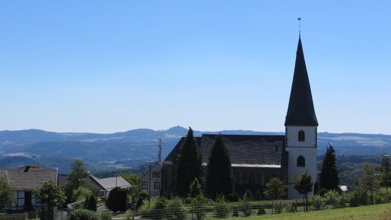 Eine Kirche mit einem hohen Turm und umgebenden grünen Wiesen. Im Hintergrund erstreckt sich eine hügelige Landschaft unter einem klaren blauen Himmel.