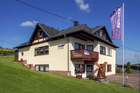 Un beau bâtiment au design moderne avec une petite terrasse en bois. Au premier plan se trouve un drapeau qui dit bienvenue.
