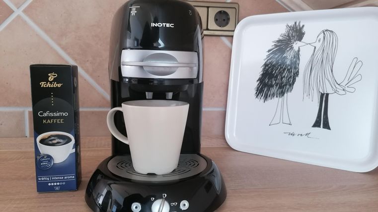 Eine Kaffeemaschine steht auf einer Arbeitsfläche, neben einer Tasse und einer Schachtel Tchibo Cafissimo Kaffee. Im Hintergrund liegt ein Tablett mit einer künstlerischen Zeichnung von Vögeln.