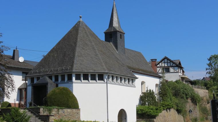 Eine charmante Kirche mit einem spitzen Turm und einem modernen, weißen Gebäude. Rundherum sind grüne Pflanzen und eine ruhige Straße.