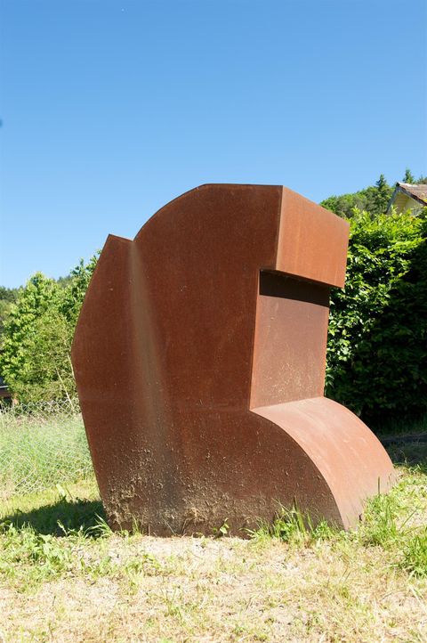 Ein modernes Kunstwerk aus rostigem Metall steht in einer grünen Landschaft. Der Himmel ist klar und blau, was die Skulptur gut zur Geltung bringt.