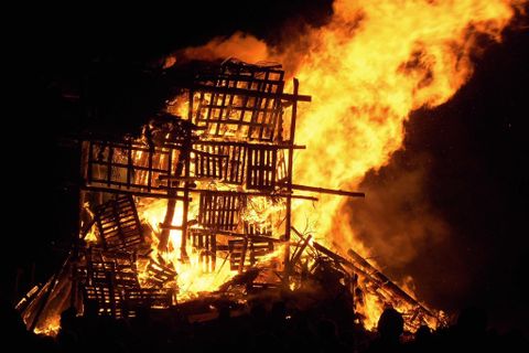 Ein großes Feuer, das einen hölzernen Stapel in Flammen setzt. Die Nacht ist erleuchtet von dem intensiven Licht des Feuers.