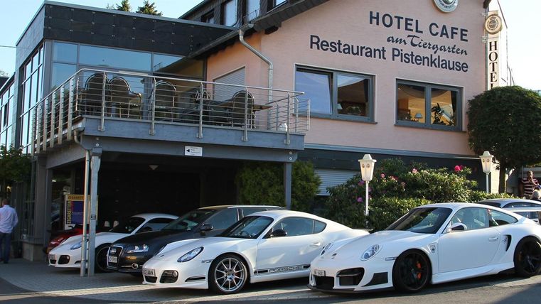 A group of elegant sports cars is parked in front of a hotel and restaurant. The buildings have an inviting atmosphere and are surrounded by green trees.