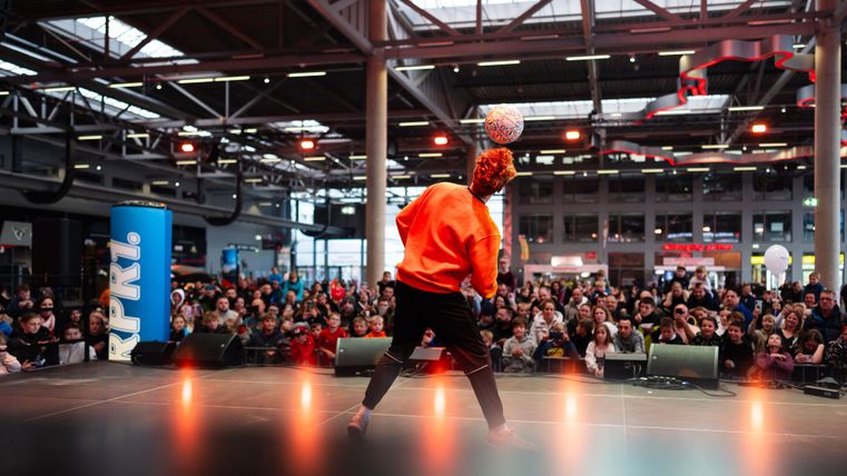 Ein Performer steht auf einer Bühne und jongliert mit einem Ball. Im Hintergrund ist eine große Menschenmenge zu sehen, die zuschaut.