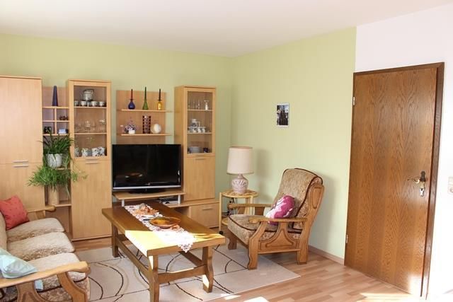 A cozy living room with a television and wooden shelves. There are comfortable armchairs and a decorative tablecloth on the table.