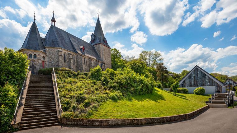 Eine beeindruckende Kirche mit Türmen steht auf einem Hügel, umgeben von grüner Landschaft. Im Hintergrund sind moderne Gebäude und ein blauer Himmel mit vielen Wolken zu sehen.