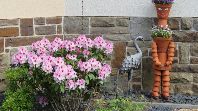 Un jardin coloré avec des fleurs roses et une figurine décorative en pots de fleurs. À côté se trouve une statue de jardin stylisée d'une cigogne.