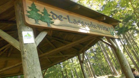 Une plaque en bois avec l'inscription "de-Lassalade-Hütte" se trouve sous une zone couverte dans la forêt. Entourée d'arbres, la scène dégage une impression de confort et de proximité avec la nature.