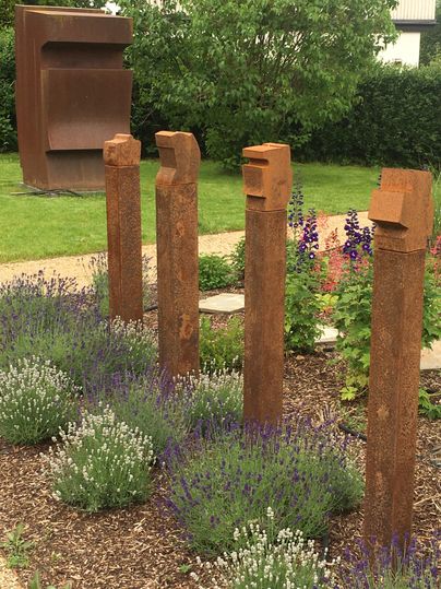 Un paysage avec quatre sculptures rouillées en forme de têtes, entourées de fleurs colorées et de lavande. En arrière-plan se dresse une grande structure couleur rouille.
