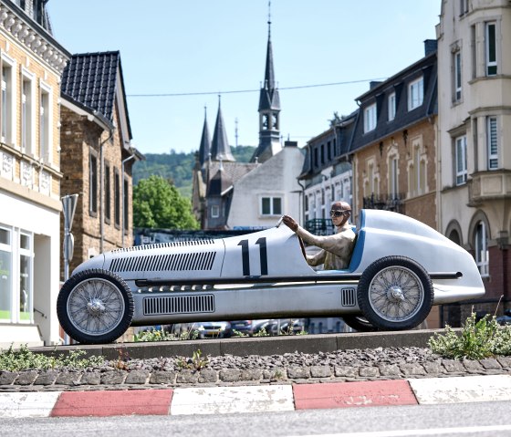 Nachbau des Silberpfeil Rennwagens mit der Nummer 11 und einem Fahrer, auf einer Verkehrsinsel mit historischen Geb&auml;uden und Kircht&uuml;rmen im Hintergrund., &copy; Stadt Adenau