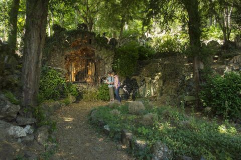 Ein malerischer Garten mit üppigem Grün und alten Steinen. Zwei Personen stehen vor einer dekorativen Nische inmitten der Natur.