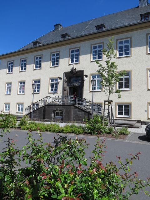 Helles Verwaltungsgebäude der Verbandsgemeindeverwaltung Adenau mit symmetrischer Fassade, mehreren Fenstern und zentralem Eingang mit Treppe, davor Pflanzen und eine Straße