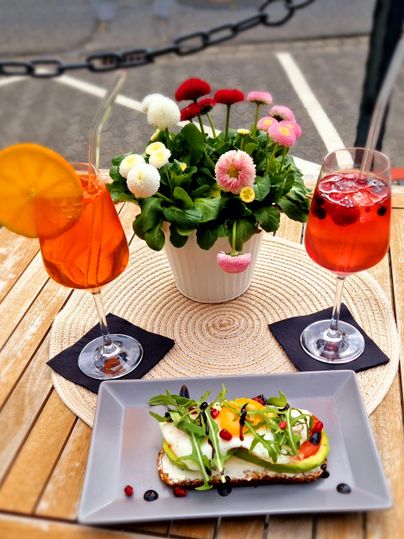 Deux cocktails colorés sont posés à côté d'une assiette avec une tartine sur une table. En arrière-plan se trouve un pot de fleurs.