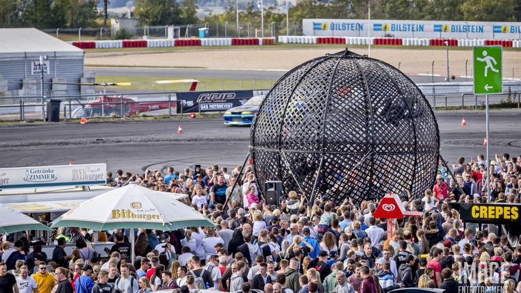 Eine große Menschenmenge versammelt sich um eine spektakuläre Attraktion auf einem Motorsportgelände. Im Hintergrund sind Autos und Verkaufsstände zu sehen.