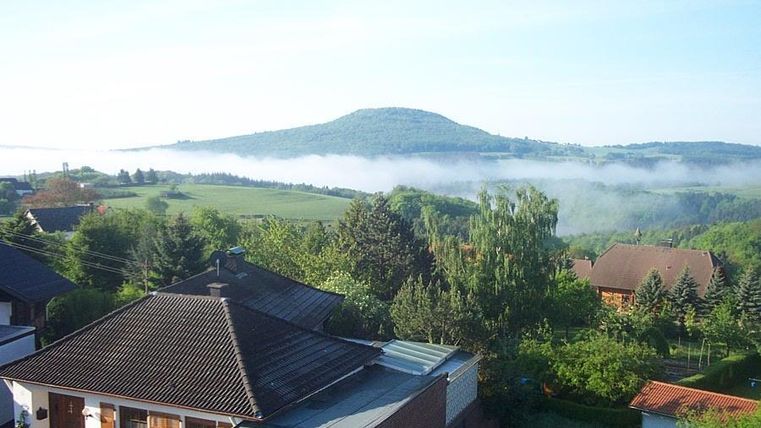 Een idylische landschap met groene heuvels en een zachte mist. Op de voorgrond zijn meerdere woonhuizen zichtbaar.