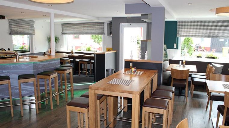 Ein modernes Restaurant mit Holzstühlen und -tischen. Der Raum hat große Fenster und ist gut beleuchtet.