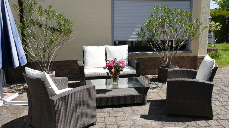 Een gezellige buitenruimte met een rieten bank en stoelen en een glazen tafel. Op de tafel staat een vaas met verse bloemen.